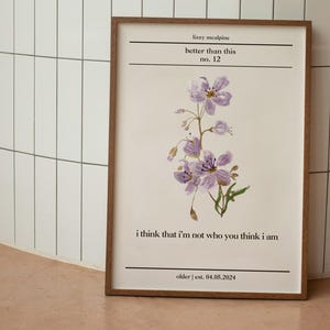 Peut inclure: Une affiche encadrée avec une illustration de fleurs violettes à l'aquarelle. Le texte "lizzy mealpine better than this no. 12" et la citation "i think that i'm not who you think i am" sont inclus. La date "older | est. 04.05.2024" est également présente.