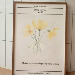Op de afbeelding: Een ingelijst drukwerk met een aquarel-illustratie van gele bloemen en de tekst "gracie m. abrams close to you no. 13 i'd give up everything to be close to you the secret of us | 06.21.2024"