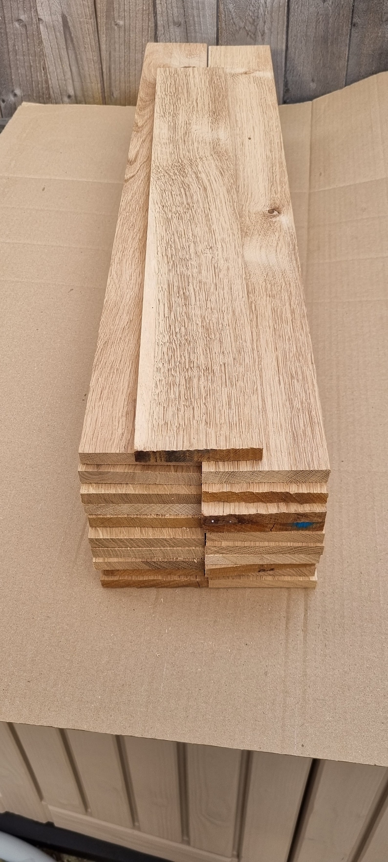 May include: A stack of 10 unfinished wooden boards, likely oak, with a natural wood grain. The boards are stacked on a brown cardboard surface.