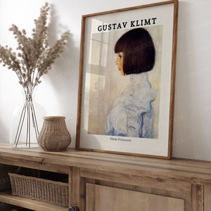 May include: A framed print of a painting by Gustav Klimt titled "Mada Primasvesi". The painting depicts a young girl with dark hair wearing a light blue dress, standing in front of a window.