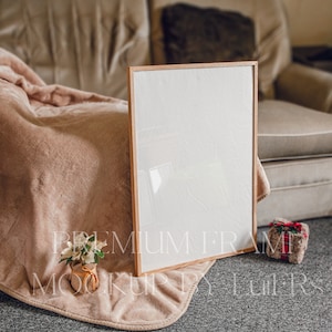 May include: A brown wooden frame with a white background sits on a beige blanket. A small potted plant with white flowers is in the foreground. A small red and black plaid gift box is on the right side of the frame.