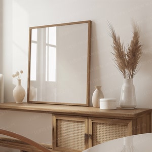May include: A square wooden picture frame rests against a white wall, above a light wood cabinet with woven cane doors and white panels. Decorative vases and a small round box are on the cabinet. A vase holds dried pampas grass.