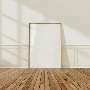 Op de afbeelding: Een houten frame met een witte achtergrond, staand op een houten vloer. Het frame bevindt zich in een kamer met witte muren en witte lambrisering. Zonlicht stroomt door een raam en werpt schaduwen op de muur.