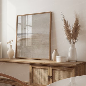 May include: A framed landscape print depicting a beach scene, set on a wooden cabinet with woven cane doors and white panels. Decorative vases and a small white box are also on the cabinet. A tall vase holds dried pampas grass.
