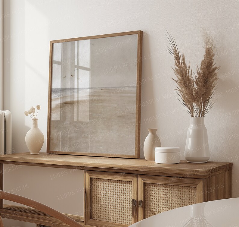 May include: A framed landscape print depicting a beach scene, set on a wooden cabinet with woven cane doors and white panels. Decorative vases and a small white box are also on the cabinet. A tall vase holds dried pampas grass.