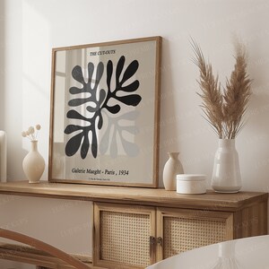 May include: A framed art print with a black and grey abstract design, labelled "THE CUT-OUTS" and "Galerie Maeght - Paris, 1934". The print is displayed on a wooden cabinet, alongside vases.