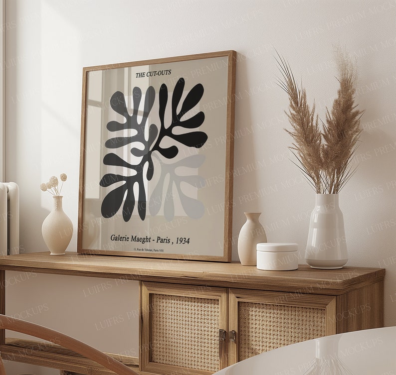 May include: A framed art print with a black and grey abstract design, labelled "THE CUT-OUTS" and "Galerie Maeght - Paris, 1934". The print is displayed on a wooden cabinet, alongside vases.