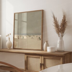 May include: A framed print of a beach scene with a wooden frame sits on a light wood cabinet. The cabinet features woven cane doors and a white panel. Decorative vases and pampas grass in a white vase complete the display.