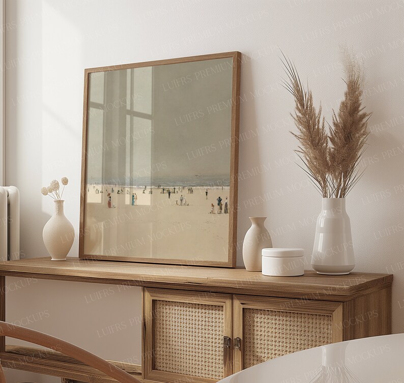 May include: A framed print of a beach scene with a wooden frame sits on a light wood cabinet. The cabinet features woven cane doors and a white panel. Decorative vases and pampas grass in a white vase complete the display.