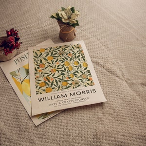 Könnte beinhalten: Ein weißer Poster mit dem Text "William Morris" in schwarzen Buchstaben. Der Poster wirbt für eine "Cotton Prints Exhibition" im "Arts & Crafts Pioneer" vom 6. Januar bis 31. Mai. Der Poster liegt auf einer beigefarbenen, strukturierten Oberfläche mit einer kleinen Topfpflanze und einem geflochtenen Korb mit roten Beeren.