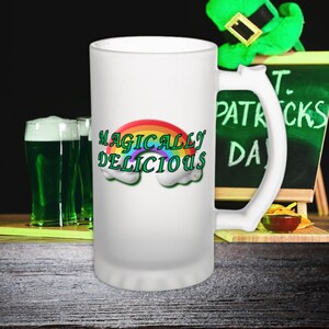 May include: White frosted glass beer stein with a rainbow and the text "Magically Delicious". The stein has a handle and is sitting on a wooden table. There is a green leprechaun hat and a chalkboard with the text "St. Patrick's Day" in the background.