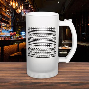 May include: A large, frosted glass beer mug with a white handle. The mug features a black and white geometric design on the front. The background shows a bar setting with dim lighting.