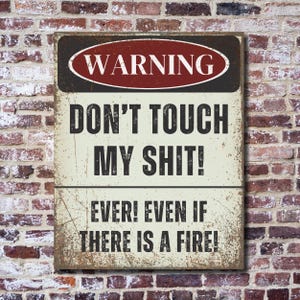 May include: A rustic metal sign with a warning sign design. The sign is white with black text and a red oval with the word "WARNING" in white text. The text on the sign reads "DON'T TOUCH MY SHIT! EVER! EVEN IF THERE IS A FIRE!"