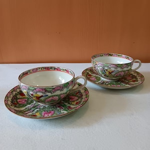 Pode incluir: Duas chávenas de chá e pires de porcelana com um desenho floral. As chávenas e os pires têm uma base branca com padrões florais rosa, verdes e amarelos. As bordas são aparadas com uma borda vermelha e dourada. As chávenas de chá têm uma pega.