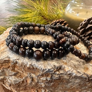May include: Three strands of dark brown wooden bead bracelets are arranged on a textured, light-colored stone. The beads are round and vary slightly in shade, creating a natural, earthy aesthetic. The bracelets are a simple, elegant accessory.
