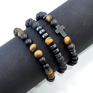 May include: Three beaded bracelets with a variety of stone beads. One bracelet features a cross charm. The beads are black, brown, and tiger's eye. The bracelets are displayed on a black surface.