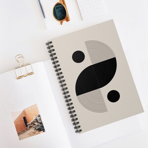 May include: A beige spiral notebook with a black and white geometric design. The design features a large black circle, a smaller black circle, and a black crescent shape with a series of concentric circles.