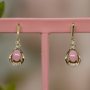 Könnte beinhalten: Ein Paar goldfarbene Ohrhänger mit einem tropfenförmigen, hellrosa Stein. Die Ohrringe haben ein dekoratives Design mit kleinen, klaren Steinen und einem Steckerverschluss. Die Ohrringe werden auf einem rosa Ständer präsentiert.