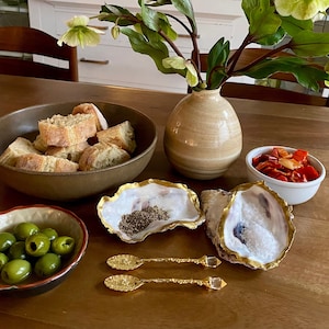 May include: A table setting with a bowl of bread, a bowl of olives, a bowl of red peppers, and two oyster shells filled with white and black grains. The table is set with a brown tablecloth and a vase of white flowers.