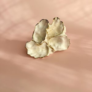 以下が含まれることがあります： 金色の縁取りが施された、オフホワイトのセラミック製の装飾皿。 花びらや貝殻の集まりのように見える、有機的で層状のデザインです。 背景はソフトピンクです。
