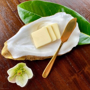 Puede incluir: Un plato decorativo con forma de concha de ostra con borde dorado, que contiene tres porciones de mantequilla. Un cuchillo de mantequilla dorado descansa junto a la mantequilla. Una hoja verde y una flor blanca y verde también están en la imagen. Sobre una superficie de madera oscura.