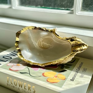 Puede incluir: Una concha de cerámica blanca con un borde dorado, sentada sobre un libro titulado "Flowers Botanical Collection No. 1980".