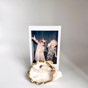 May include: A white seashell with a gold-painted rim, next to a white framed photograph of two children. The photograph shows two children, one wearing sunglasses and the other wearing a patterned shirt.