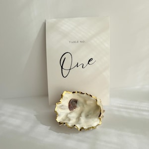 Könnte beinhalten: Eine natürliche Austernschale mit goldfarbenem Rand liegt vor einer weißen Karte. Auf der Karte steht "TABLE NO. One" in eleganter schwarzer Schrift. Die Schale ist cremefarben mit einem Hauch von Lila.