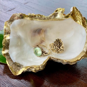 May include: A white oyster shell with gold leaf accents, used as a ring dish. Two gold rings are displayed in the shell. One ring has a large green gemstone.
