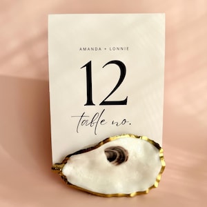 Peut inclure: Une carte de numéro de table blanche avec les noms "Amanda + Lonnie" et le numéro "12" en noir. La carte repose sur une coquille d'huître blanche avec un bord doré. Les mots "table no." sont écrits en script.
