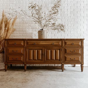 May include: A long wooden sideboard featuring a warm brown finish, six drawers with brass-toned hardware, and two central cabinet doors with vertical panel detailing. The piece stands on four tapered legs and is topped with a ceramic crock.