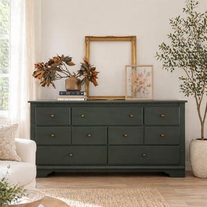 May include: A dark green dresser with eight drawers and gold knobs. The dresser is topped with a gold framed mirror, a vase with dried foliage, and a framed floral print. A white armchair and a potted plant are visible.