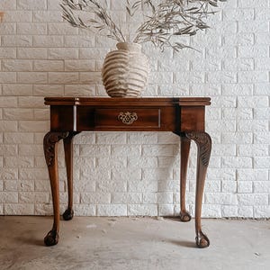 May include: A dark brown antique table with a single drawer and ornate hardware. The table has curved legs with claw feet. A textured vase with dried branches sits on top. The table is approximately 30 inches long.