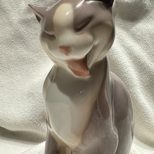 Könnte beinhalten: Eine Keramikfigur eines grau-weißen Katers mit einem verspielten Ausdruck. Die Katze sitzt mit geneigtem Kopf und leicht geöffnetem Maul, als würde sie gleich miauen.