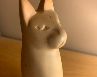 May include: A cream-colored, carved figurine of a dog with pointed ears. The sculpture has a smooth finish with subtle variations in color and texture. The dog's features are simplified, with a prominent snout and alert expression. The figurine is set against a neutral background.