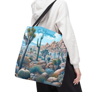 Könnte beinhalten: Eine Tragetasche mit einer Wüstenlandschaft mit Kakteen, Joshua-Bäumen und felsigem Gelände unter blauem Himmel. Das Wort "California" ist in einer Schriftart geschrieben. Die Tasche hat einen schwarzen Schulterriemen.