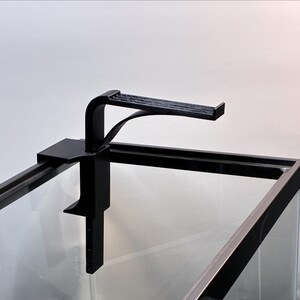 May include: A black metal aquarium light fixture is clamped onto the edge of a glass aquarium. The fixture has a curved arm and a rectangular light support. The aquarium is empty, with a clear glass front.