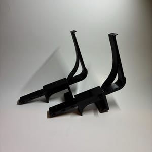 May include: Two black, angular, 3D-printed objects. Each has a rectangular base with several holes and a curved, upward-reaching support. The supports have a hook-like shape at the top. The objects are set against a white background.