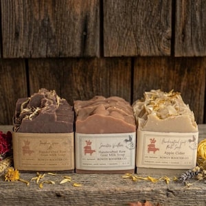 May include: Three rectangular bars of Rowdy Rooster Co. handcrafted raw goat milk soap. The left bar is dark brown, the center is light brown, and the right is beige with dried petals. Each has a paper label with text and a rooster logo.