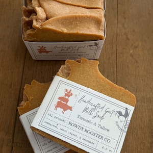 May include: Three rectangular bars of golden-brown turmeric and tallow goat milk soap. Each bar features a white paper label with the text Handcrafted Goat Milk Soap, Turmeric & Tallow, ROWDY ROOSTER CO., all natural, 4.3 oz.