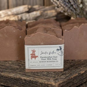 May include: Several rectangular bars of brown goat milk soap with a textured top. A white paper label wraps around the front bar, featuring the text Sweater Weather, Handcrafted Raw Goat Milk Soap, Rowdy Rooster Co., and 4.3 oz.