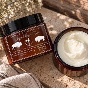 Puede incluir: Dos frascos de vidrio ámbar que contienen sebo de bisonte batido Rowdy Rooster Co. Un frasco está cerrado con una tapa negra y una etiqueta que dice Rowdy Rooster Co. Whipped Bison Tallow, All Natural, 100% Handmade, For Face & Body, Unscented, 113 g.