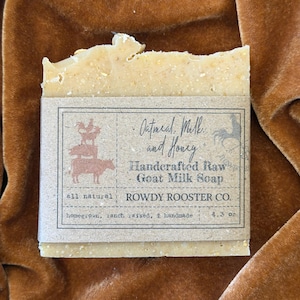 May include: A rectangular bar of tan, textured soap wrapped in a brown paper label. The label features a stack of farm animals, the text Oatmeal, Milk and Honey, Handcrafted Raw Goat Milk Soap, Rowdy Rooster Co., and the weight 4.3 oz.