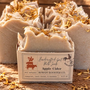 May include: Three bars of beige goat milk soap topped with dried yellow flower petals. The front bar features a paper label reading Handcrafted Goat Milk Soap Apple Cider, all natural, ROWDY ROOSTER CO., homegrown, ranch raised, & handmade, 4.3 oz.