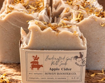 Könnte beinhalten: Drei Stücke beigefarbene Ziegenmilchseife, garniert mit getrockneten gelben Blütenblättern. Das vordere Stück hat ein Papieretikett mit der Aufschrift Handcrafted Goat Milk Soap Apple Cider, all natural, ROWDY ROOSTER CO., homegrown, ranch raised, & handmade, 122 g.