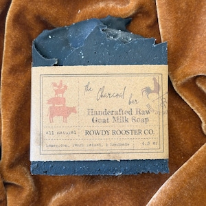 May include: A rectangular black charcoal bar of handcrafted raw goat milk soap wrapped in a tan paper label. The label features a rooster, pig, and cow illustration, the text The Charcoal bar, Handcrafted Raw Goat Milk Soap, ROWDY ROOSTER CO., and 4.3 oz.
