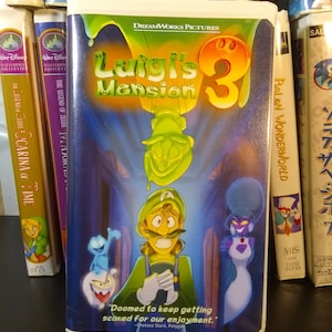 May include: A VHS tape for the video game "Luigi's Mansion 3" featuring Luigi in a green outfit, surrounded by cartoon ghosts. The cover has the DreamWorks Pictures logo and the quote "Doomed to keep getting scared for our enjoyment."