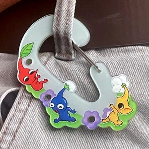 Könnte beinhalten: Ein hellblauer Karabinerhaken mit drei Cartoon-Pikmin-Charakteren in Rot, Blau und Gelb. Der Haken ist mit lila Blumen und grünem Gras verziert. Der Haken ist an einer Gürtelschlaufe befestigt.