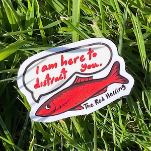 May include: A sticker with a red fish design and the text "I am here to distract you." in a speech bubble. The text "The Red Herring" is below the fish. The sticker is set against a backdrop of green grass.