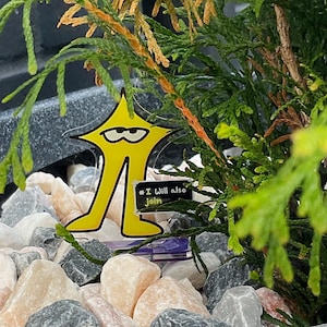 May include: A yellow star-shaped ornament with a cartoon face and legs, hanging in a small evergreen tree. The ornament has the text "# I Will also Join" on a small black rectangle. The tree is surrounded by small rocks.